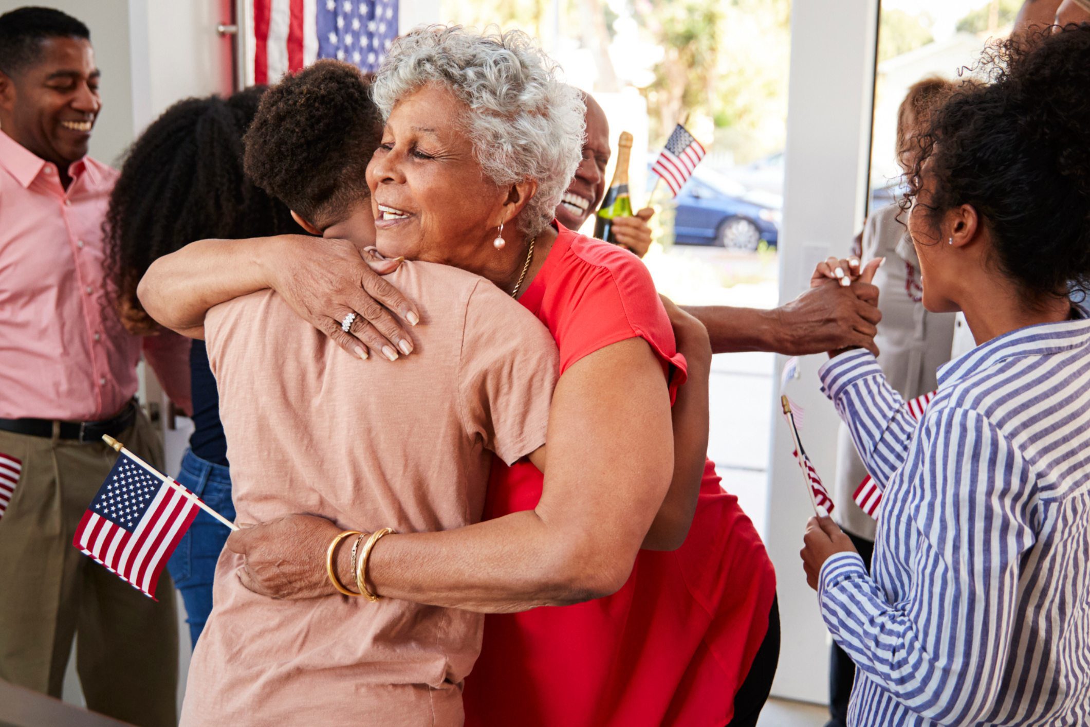 Two women warmly embracing outdoors with an American flag in the background.